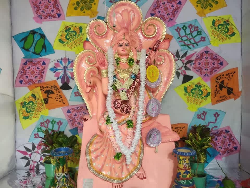 Pink Saraswati idol with garlands, set against a backdrop of colourful kite decorations